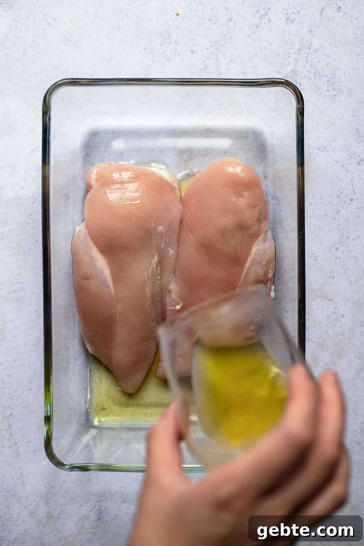 two raw chicken breasts in a baking dish with oil being poured over them