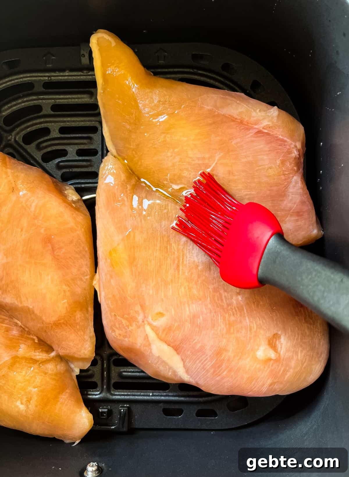 A brush applying olive oil to the top of the chicken breasts in the air fryer basket.