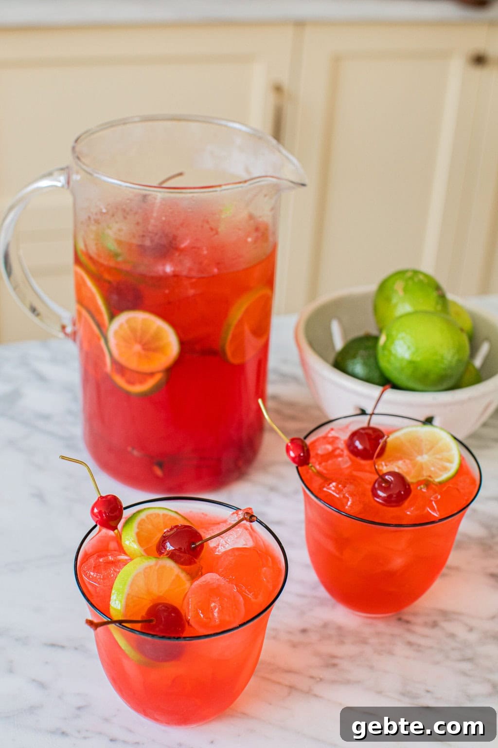 a large pitcher of dirty shirley cocktails beside two cocktail glasses full of the drink and a small bowl of extra limes