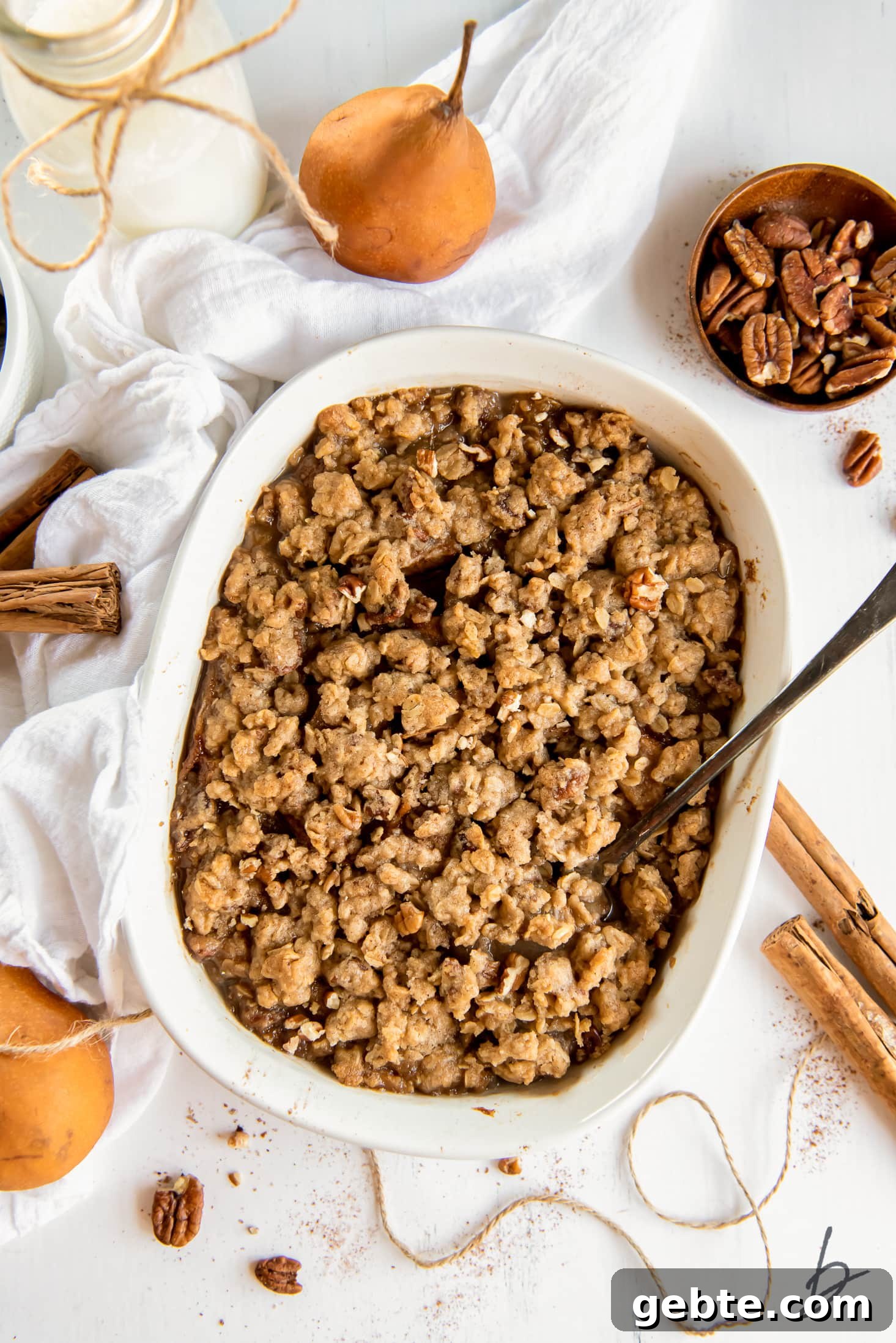 Pear crisp in a baking dish next to white cloth, pears and cinnamon sticks.