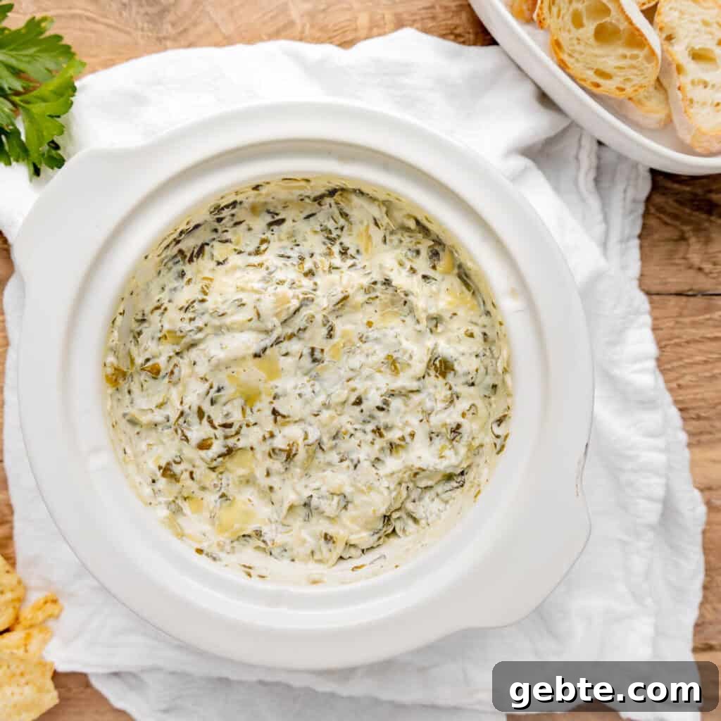 A white dish filled with creamy spinach and artichoke dip sits on a wooden table with a white cloth. Slices of bread are next to the dish.