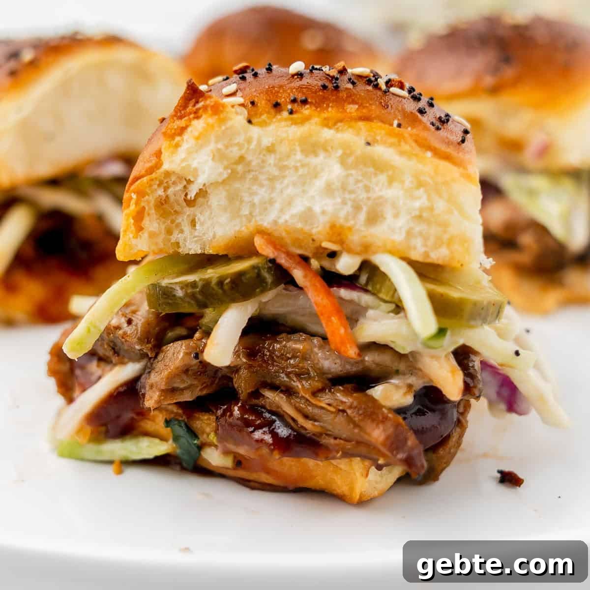 A pulled pork slider with pickles, coleslaw, and barbecue sauce on a sesame-topped bun, displayed on a white plate.
