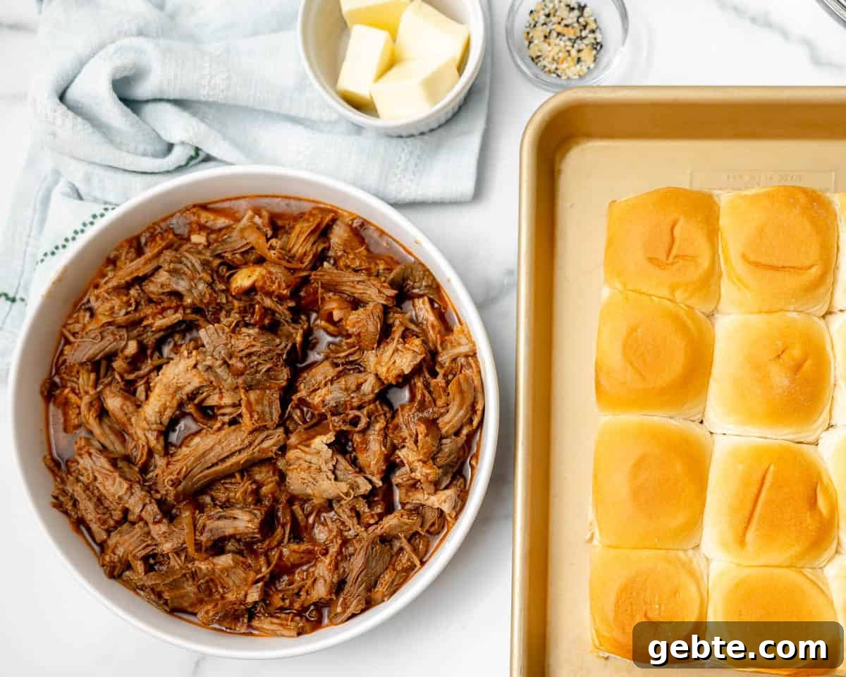 A bowl of shredded pork in sauce next to a tray of slider buns, with a small bowl of butter cubes and a dish of seasoning on a white surface.