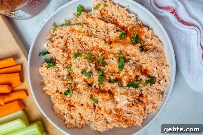 Shredded chicken with a creamy, spicy buffalo sauce in white bowl next to carrots and celery