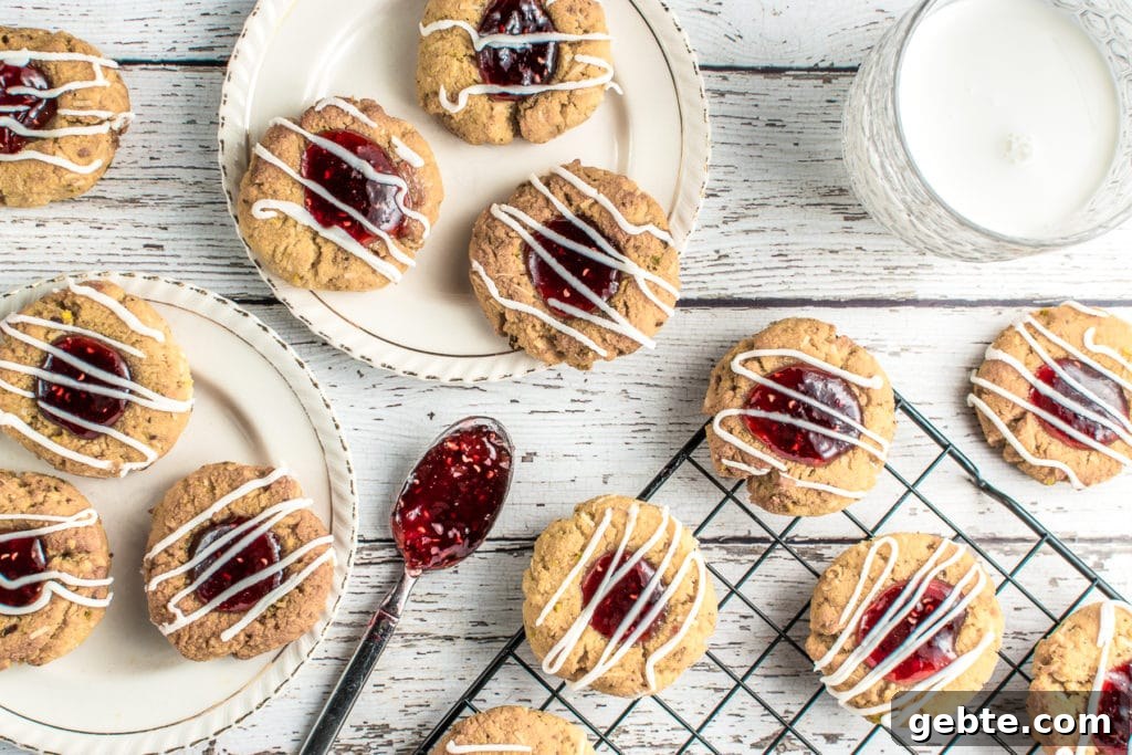 Keto raspberry jam thumbprint cookies on two, ivory, vintage dessert plates set on a rustic white barnyard backdrop. A glass of milk and a cooling rack with more cookies, along with a teaspoon filled with gooey, raspberry jam, complete the picture.