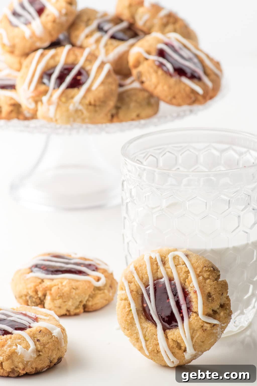 Set on a white backdrop are three, almond flour thumbprint cookies in the foreground, with one propped against a glass half filled with milk. The background contains a blurred, pedestal cake plate piled with raspberry jam-filled, almond flour, thumbprint cookies with pistachios.  All cookies are drizzled with white icing.