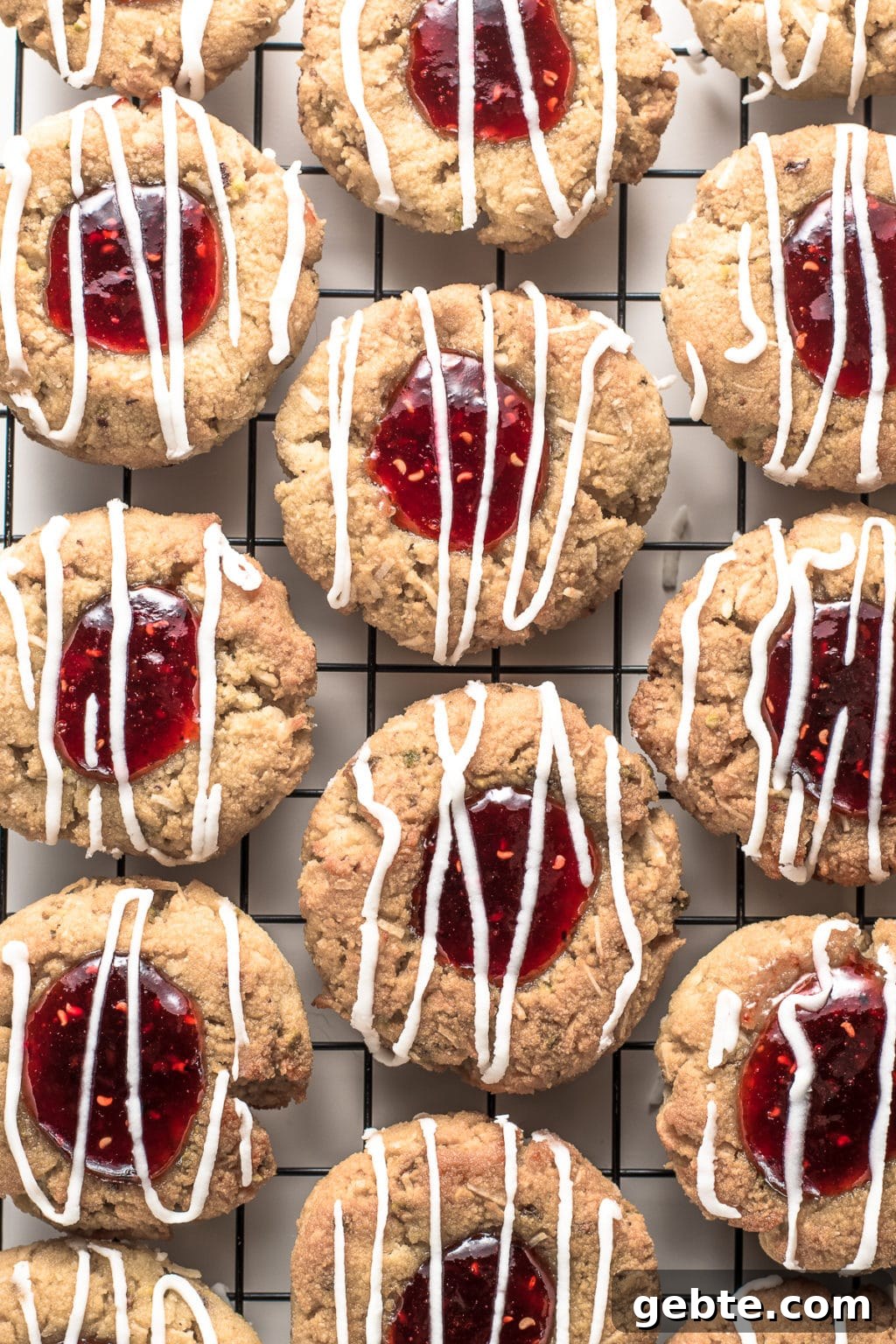 Resting on a white backdrop, is a metal cookie rack covered with raspberry jam-filled, thumbprint cookies drizzled with sugar-free, white glaze.