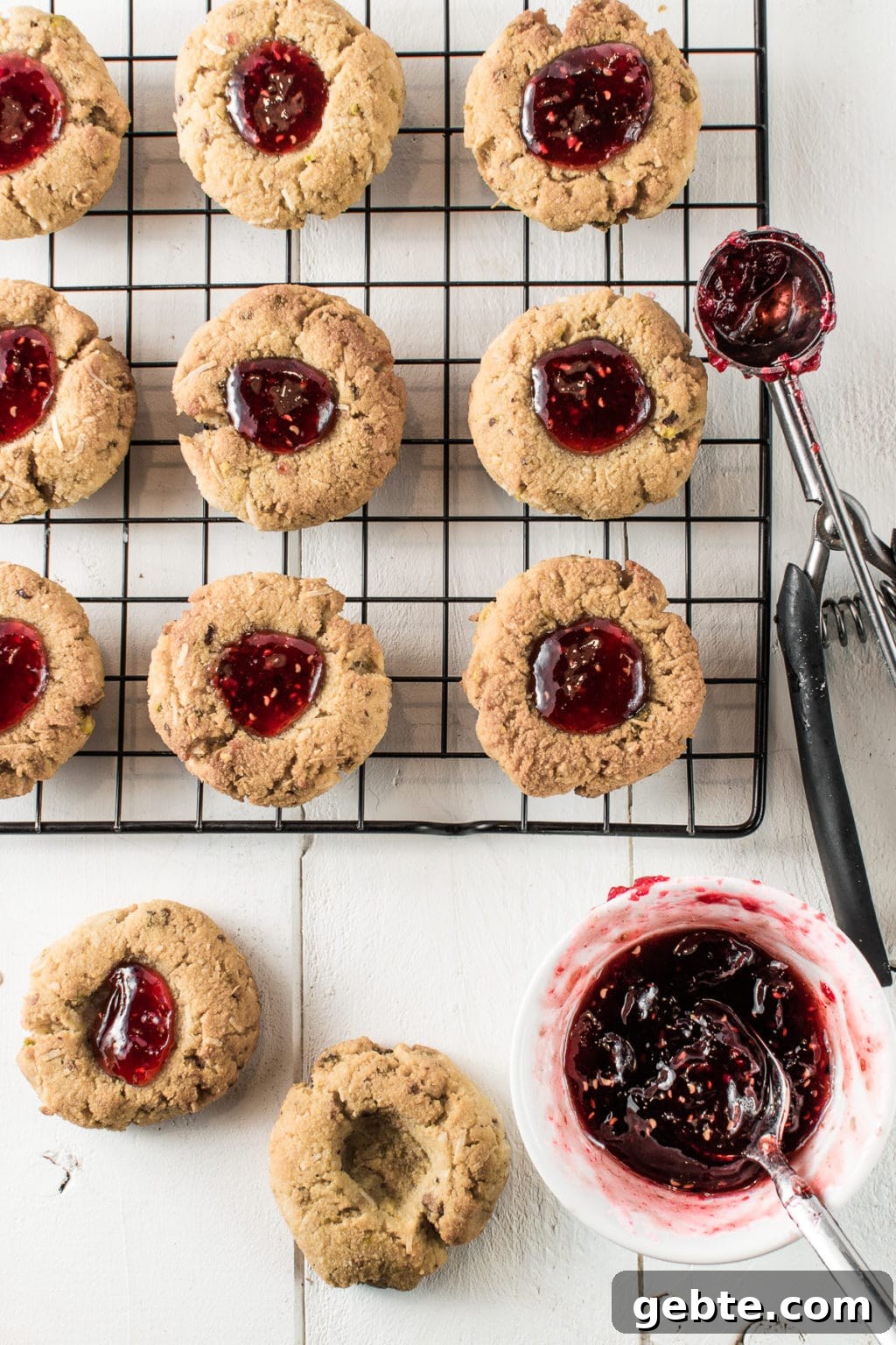 Set on a white backdrop, are nine thumbprint cookies filled with gooey, raspberry jam spaced evenly on a cooling rack. A stainless cookie scoop, half full of sticky jam, rests on the right side of the metal rack.  Below the rack, are two cookies--one filled with jam, and one with an empty divot. Beside the empty cookie is a jam-filled teaspoon inside a small, white bowl filled with sticky, raspberry jam.