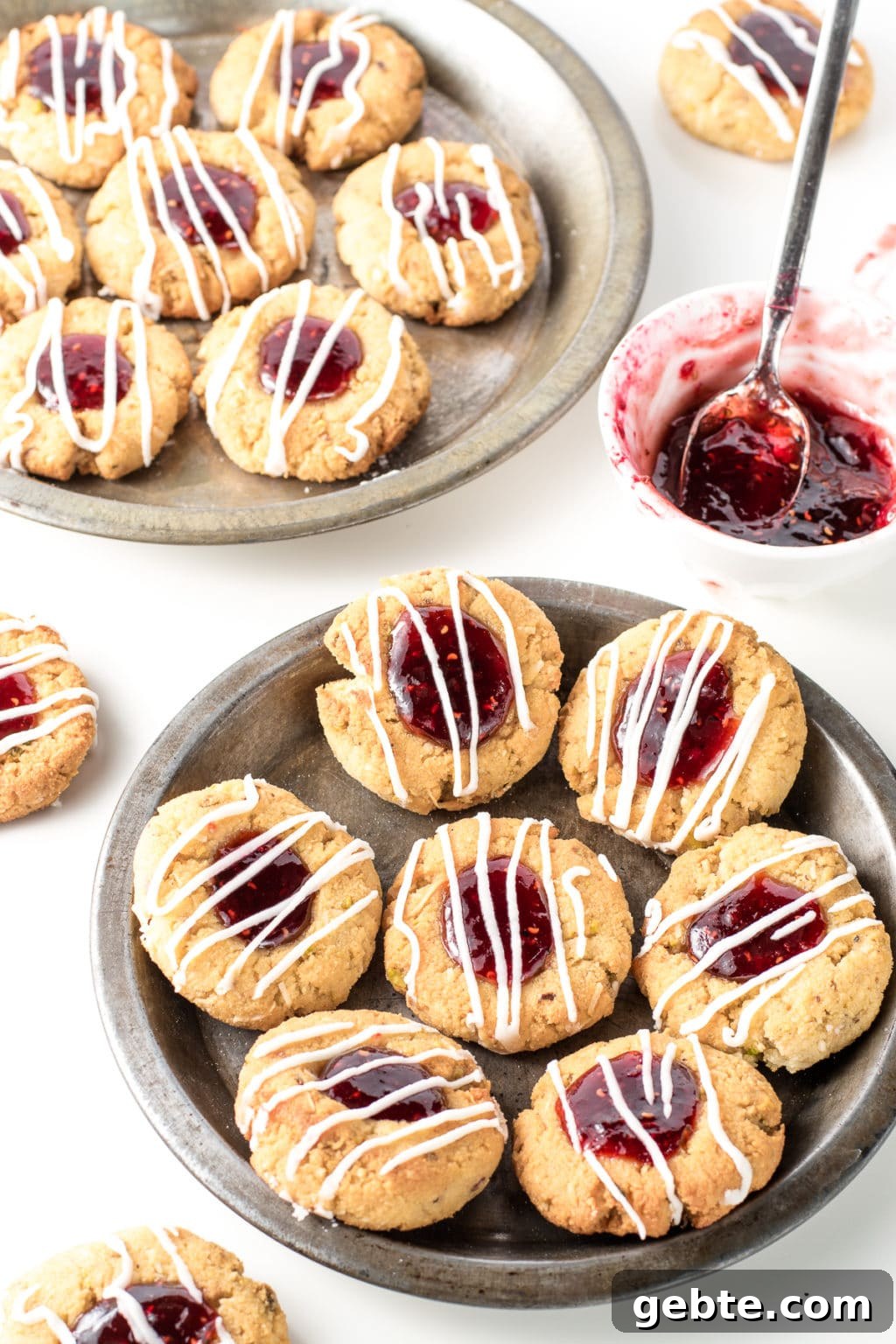 Two metal pie plates filled with thumbprint cookies. There are two cookies resting directly on the white background. To the right of centre is a small, white bowl with a teaspoon in it.  They are filled with gooey, raspberry jam.