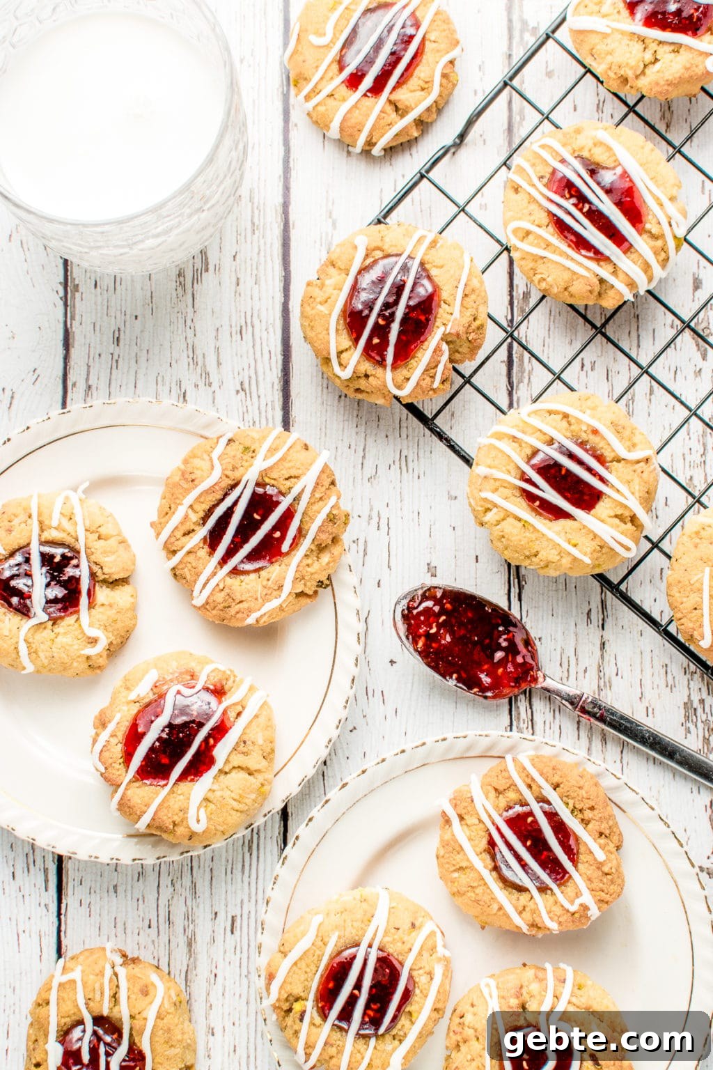 Rustic white barnyard backdrop with a glass of milk on the top left side and a thumbprint cookie beside it to the right. Centre right is the corner of a cooling rack placed diagonally filled with cookies. Bottom left has a vintage white plate with gold trim on which rests three cookies. Beside the plate is a jam-filled teaspoon resting on the backdrop.