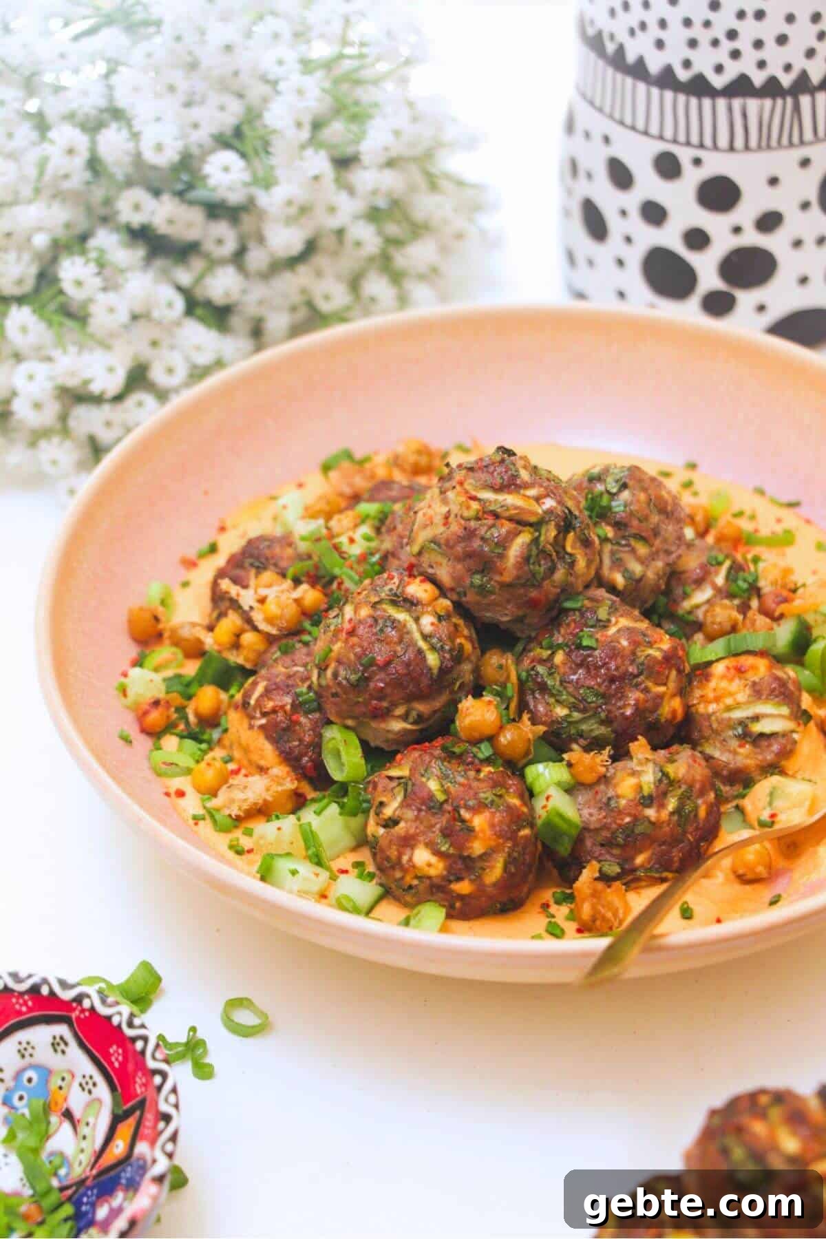 Meatballs piled on top of smoky red pepper sauce with flowers in the background.