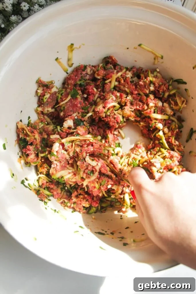 Hand holding a spoon mixing zucchini, feta meatball mixture.