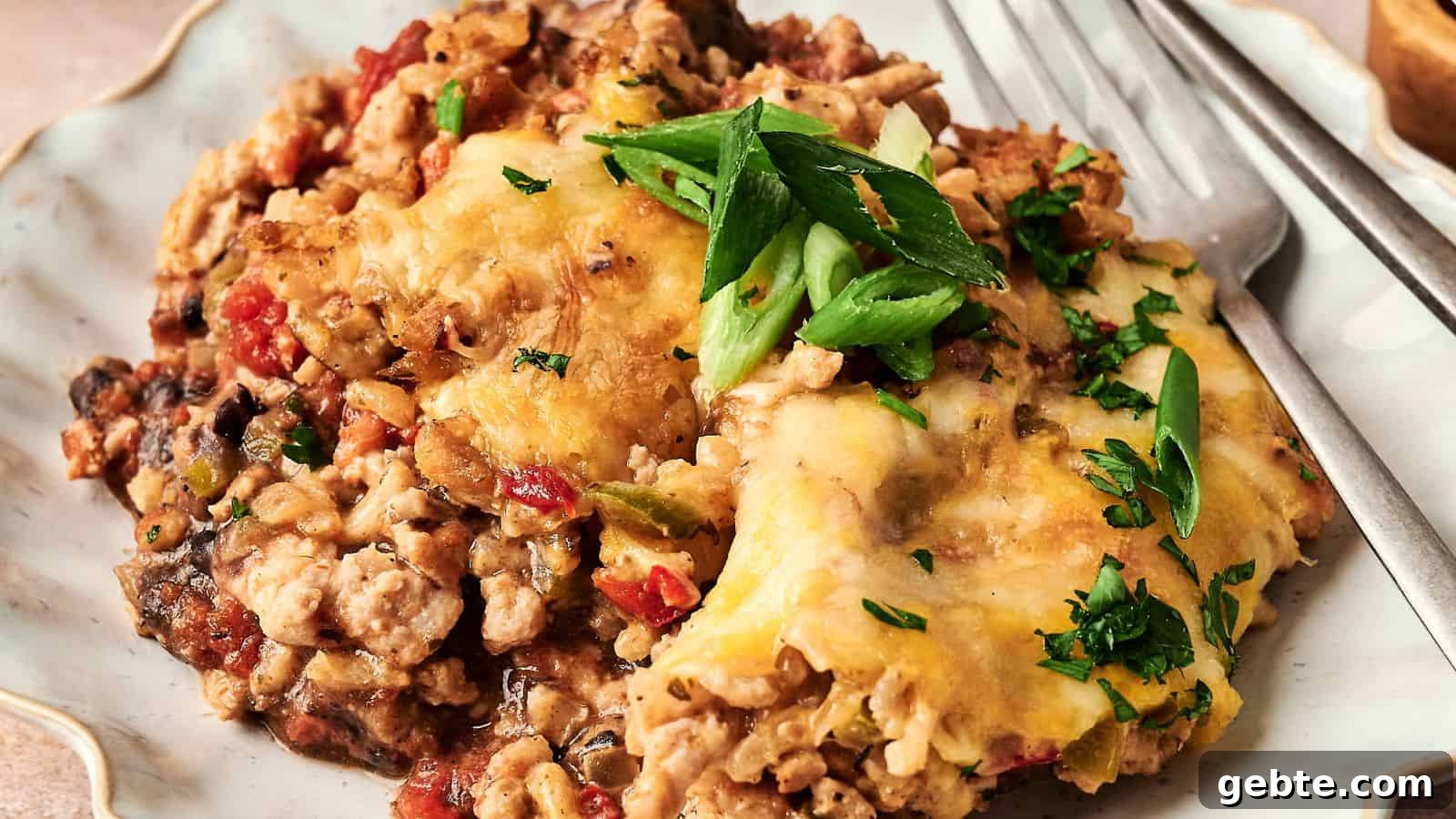 A plated serving of cheesy casserole with ground meat, topped with sliced green onions and parsley, accompanied by a fork.