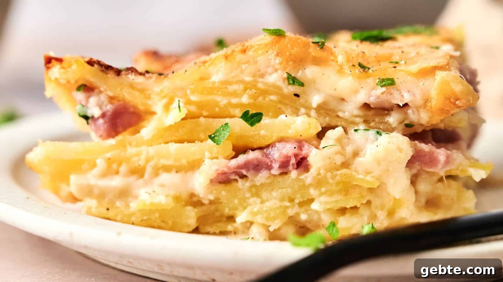 A close-up of a slice of potato casserole with layers of potatoes, ham, and creamy sauce, garnished with chopped parsley on a plate.