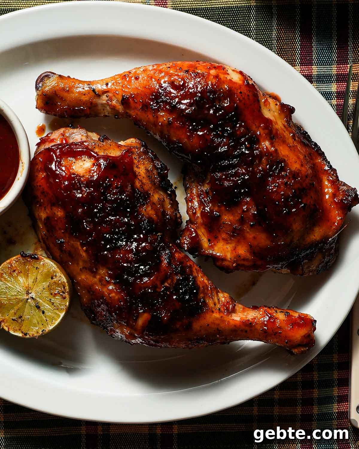 A white platter with 2 cooked chicken legs with bbq sauce, grilled lime and a side of barbecue sauce.