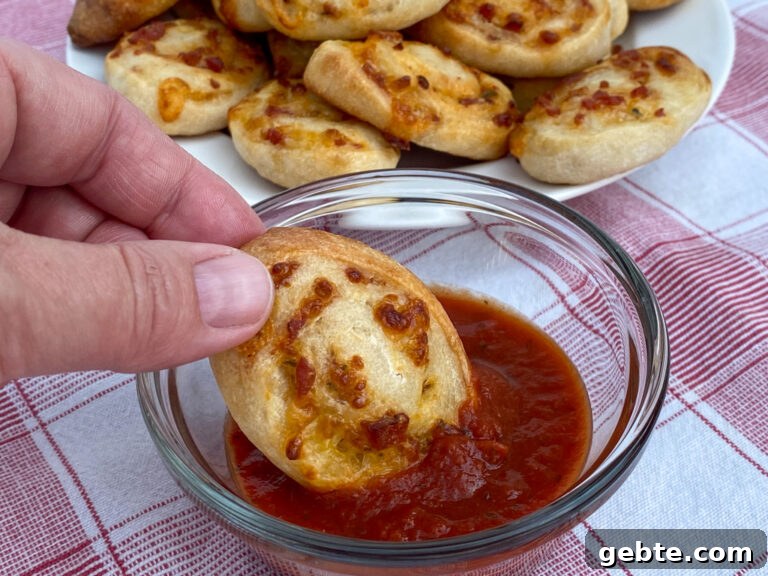 Hand dipping a pinwheel in a small glass dish with pizza sauce on a red and white plaid napkin with a plate full in the background.