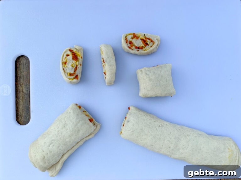 Roll of refrigerated pizza dough filled with chopped pepperoni and cheese, then rolled up and being cut into slices to make pin wheels.