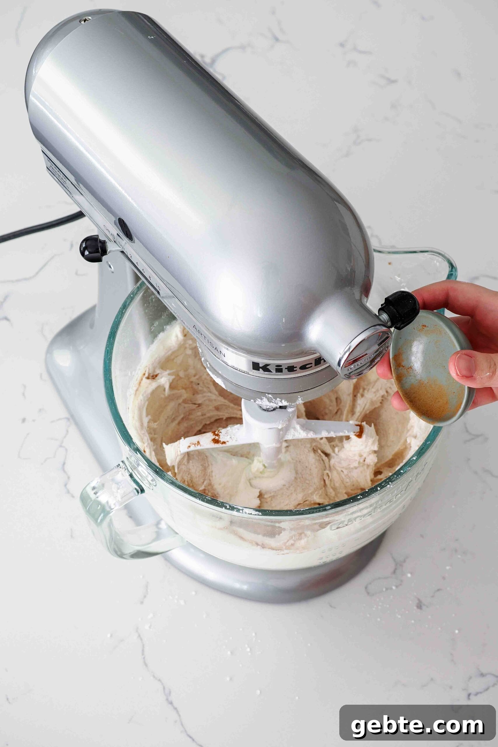 A hand adds ground cinnamon to buttercream in a stand mixer bowl.