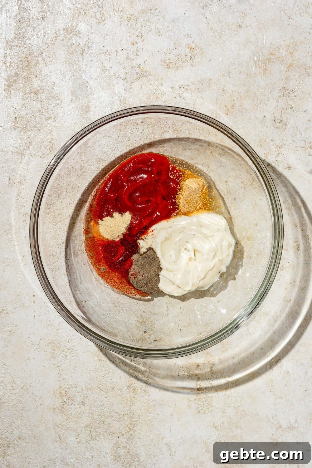 A glass bowl with ingredients to make copycat Zaxby's sauce including ketchup, ranch dressing, vinegar, onion, and seasonings. The bowl is on a tan counter.