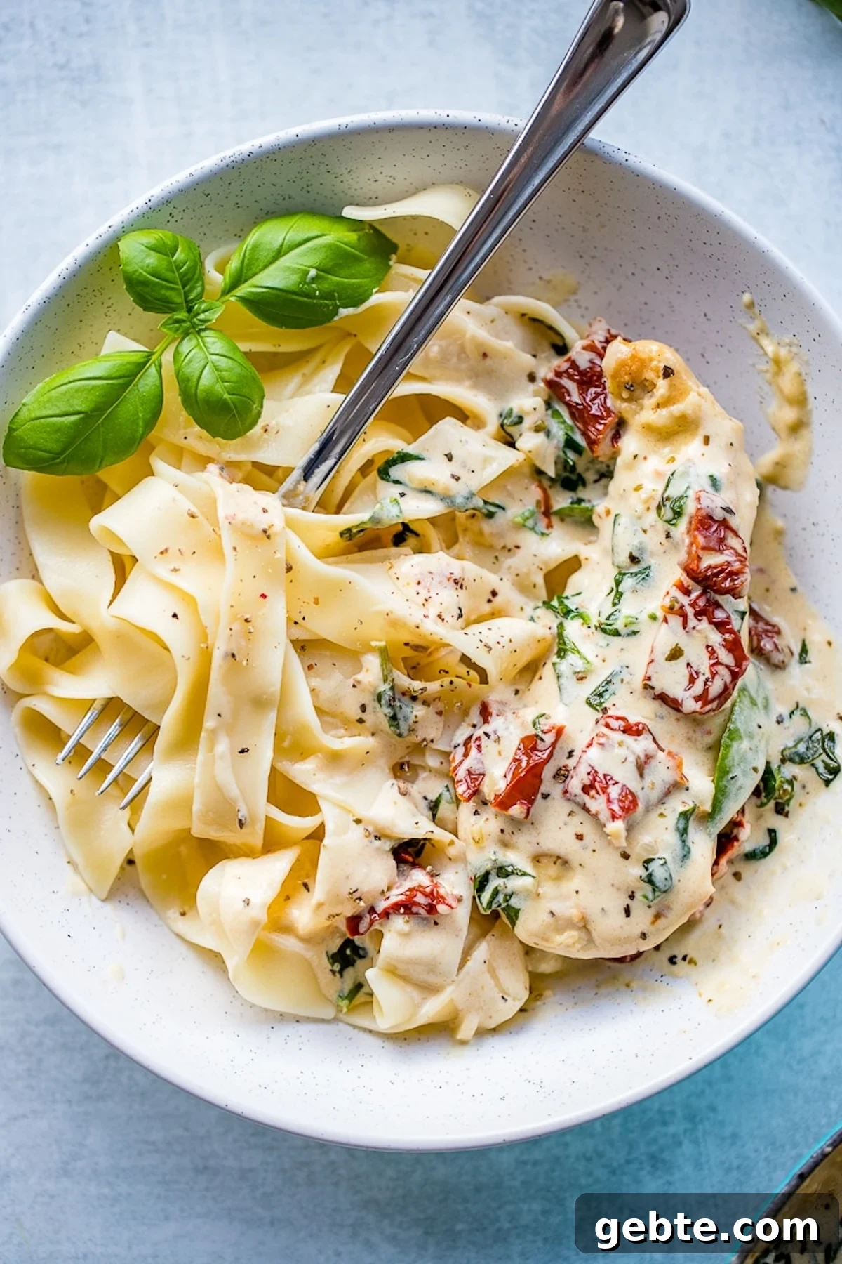 A serving bowl filled with ribbon pasta that's been tossed in a white sun-dried tomato  and spinach sauce. Fresh basil sits on the side.