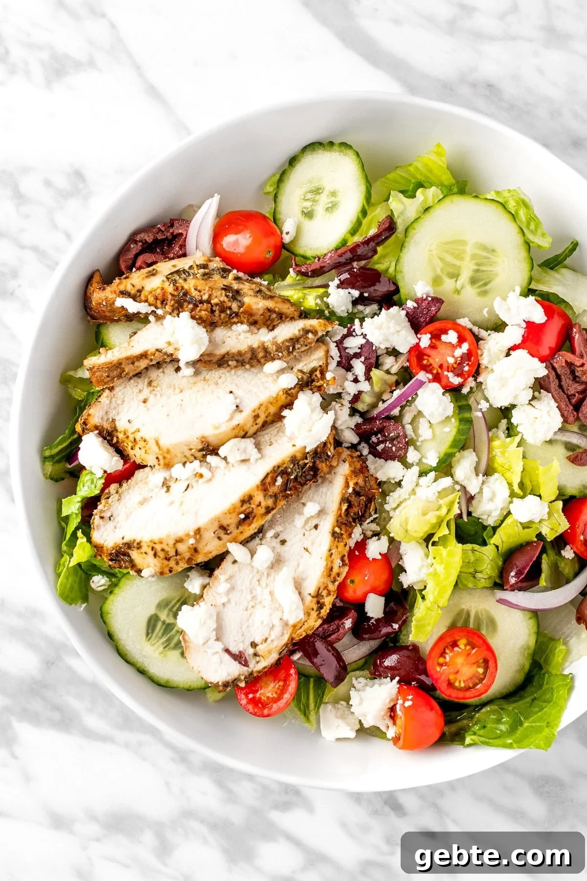 A white salad bowl is filled with roughly chopped lettuce that's topped with slice chicken Brest, sliced cucumber, halved cherry tomatoes, sliced red onion, Kalamata olives, and crumbled feta cheese.