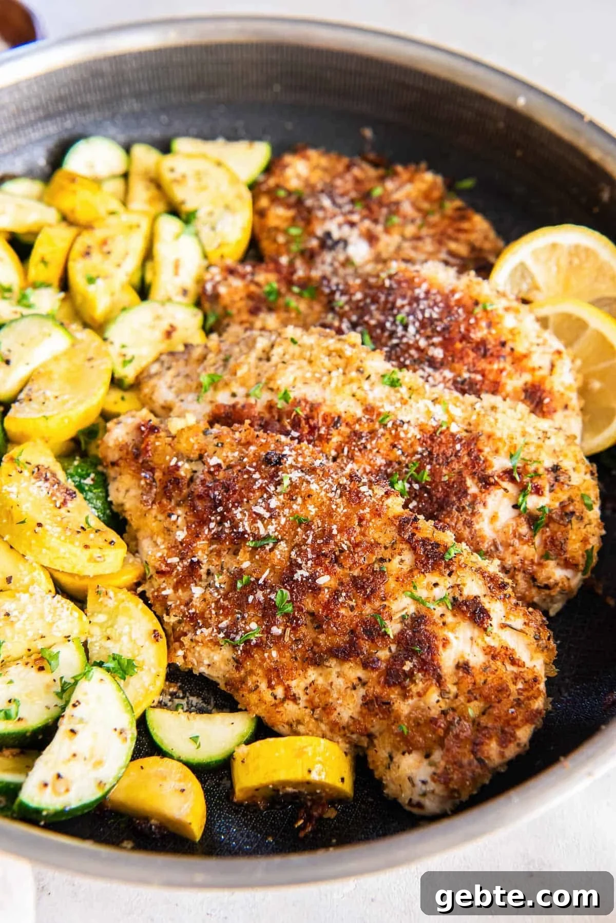A large skillet has cooked chicken breast that's coated in a golden brown parmesan breading, next to sautéed squash and zucchini moons.
