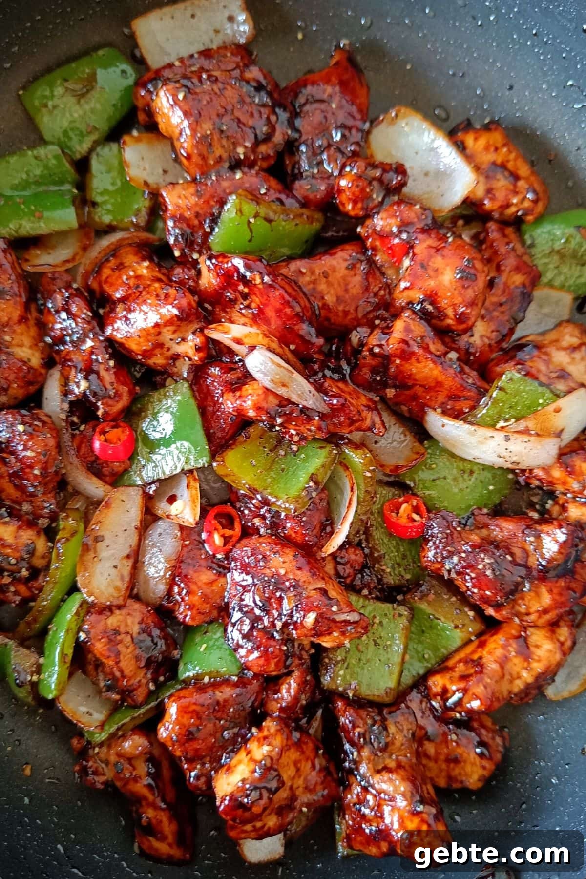 Chunks of chicken coated in a deep red glaze in a skillet with largely diced green bell peppers, white onion, and red hot peppers.