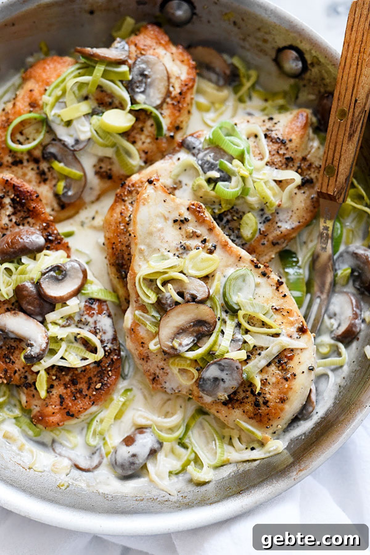 A stainless steel skillet is filled with seared chicken breasts that are topped with sliced mushrooms and leeks. They are sitting in a creamy white sauce with additional mushrooms and leeks.