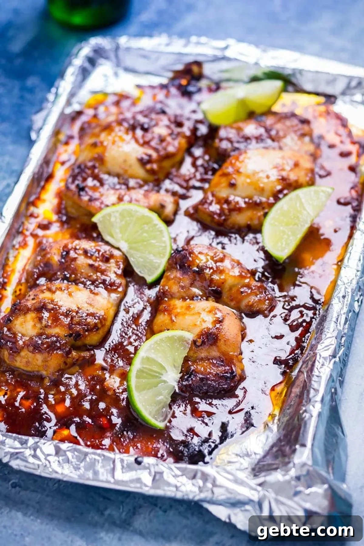 A sheet pan coated in aluminum foil is full of of chicken that's coated in and swimming in a red honey chipotle sauce. Slices of lime are next to each chicken thigh.