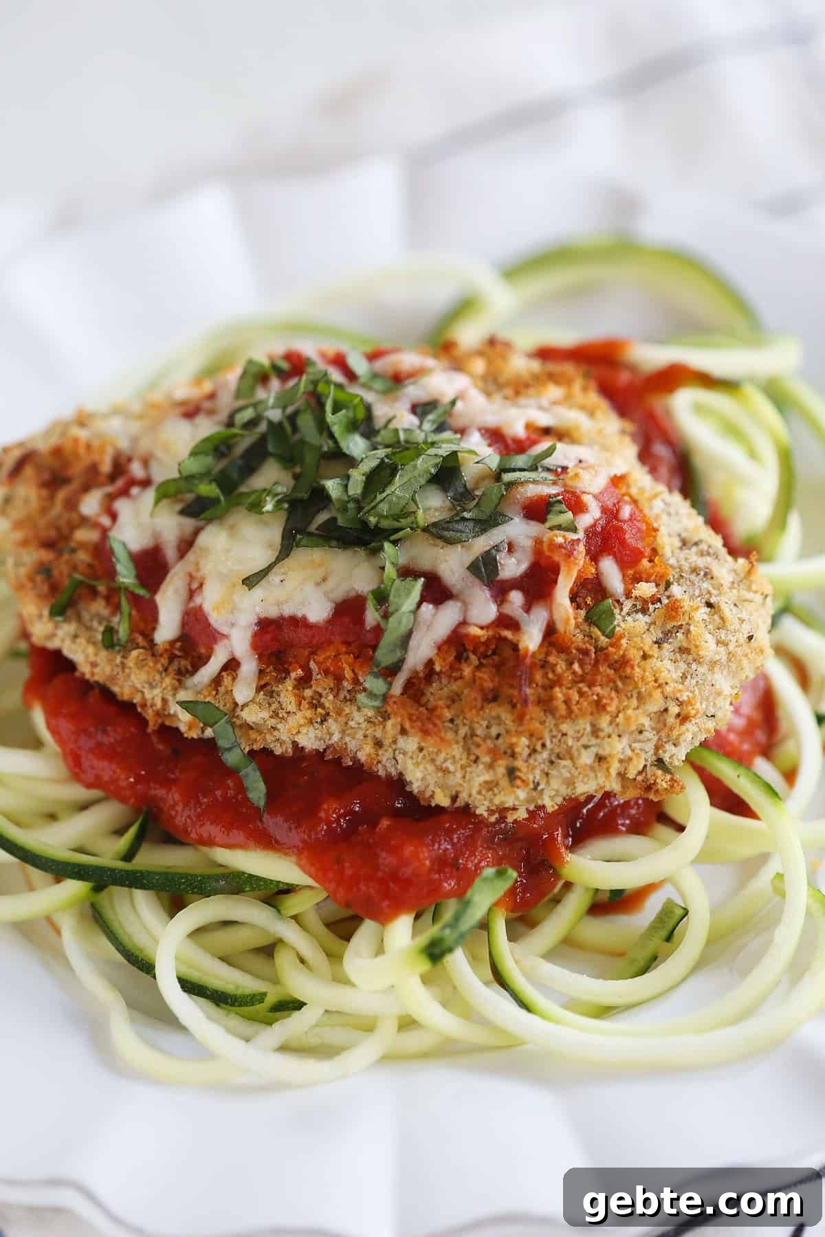 A breaded chicken breast is topped with res sauce and melted white cheese. It sits on a bed of zucchini noodles on a white dinner plate.