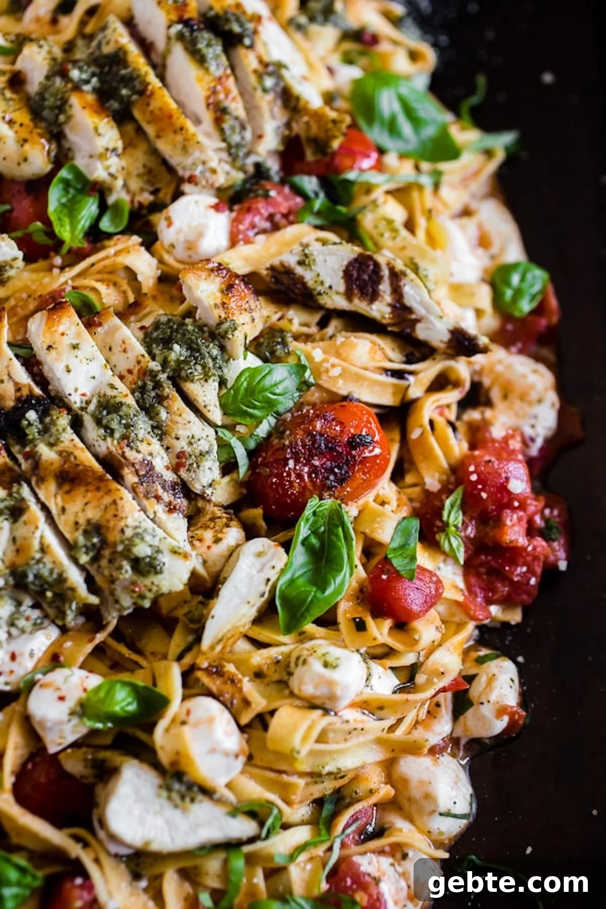 Ribbon pasta topped with seared cherry tomatoes, fresh basil, mozzarella pearls, and sliced chicken breast on top of a black serving plate.