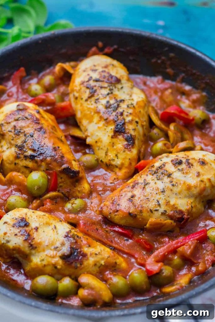 Seared chicken breasts on top of a red sauce with green olives and red peppers, all in a cast iron skillet.