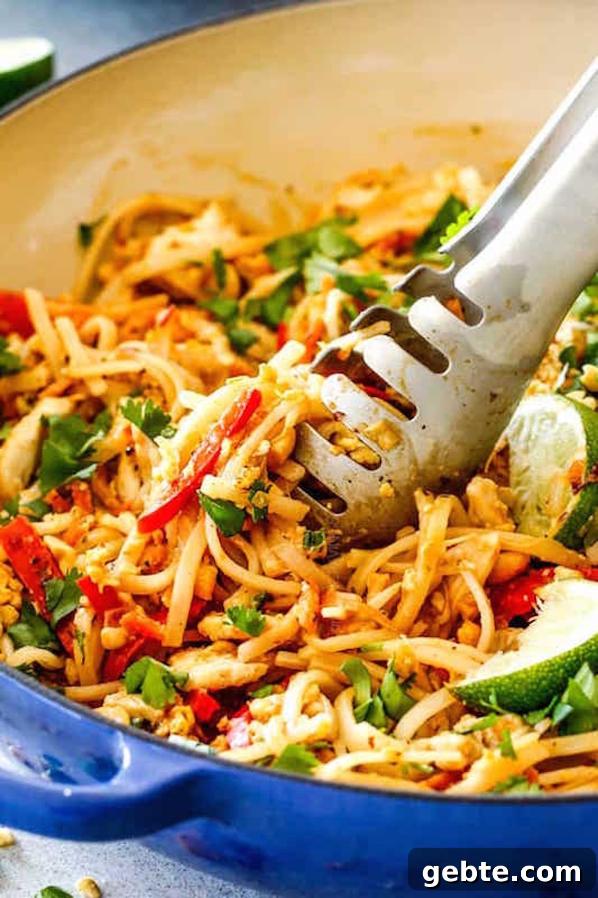 A large skillet filled with a chicken pad Thai that as visible chopped herbs, red peppers, and lime slices.