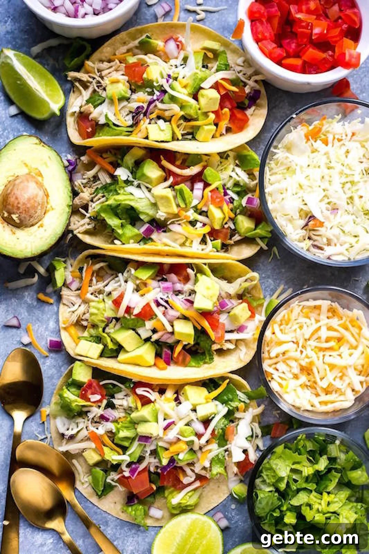 Four corn tortilla tacos generously filled with shredded chicken, chopped avocado, chopped lettuce, and diced red onion sitting next to two bowls of cheese, lime wedges, and a half avocado.