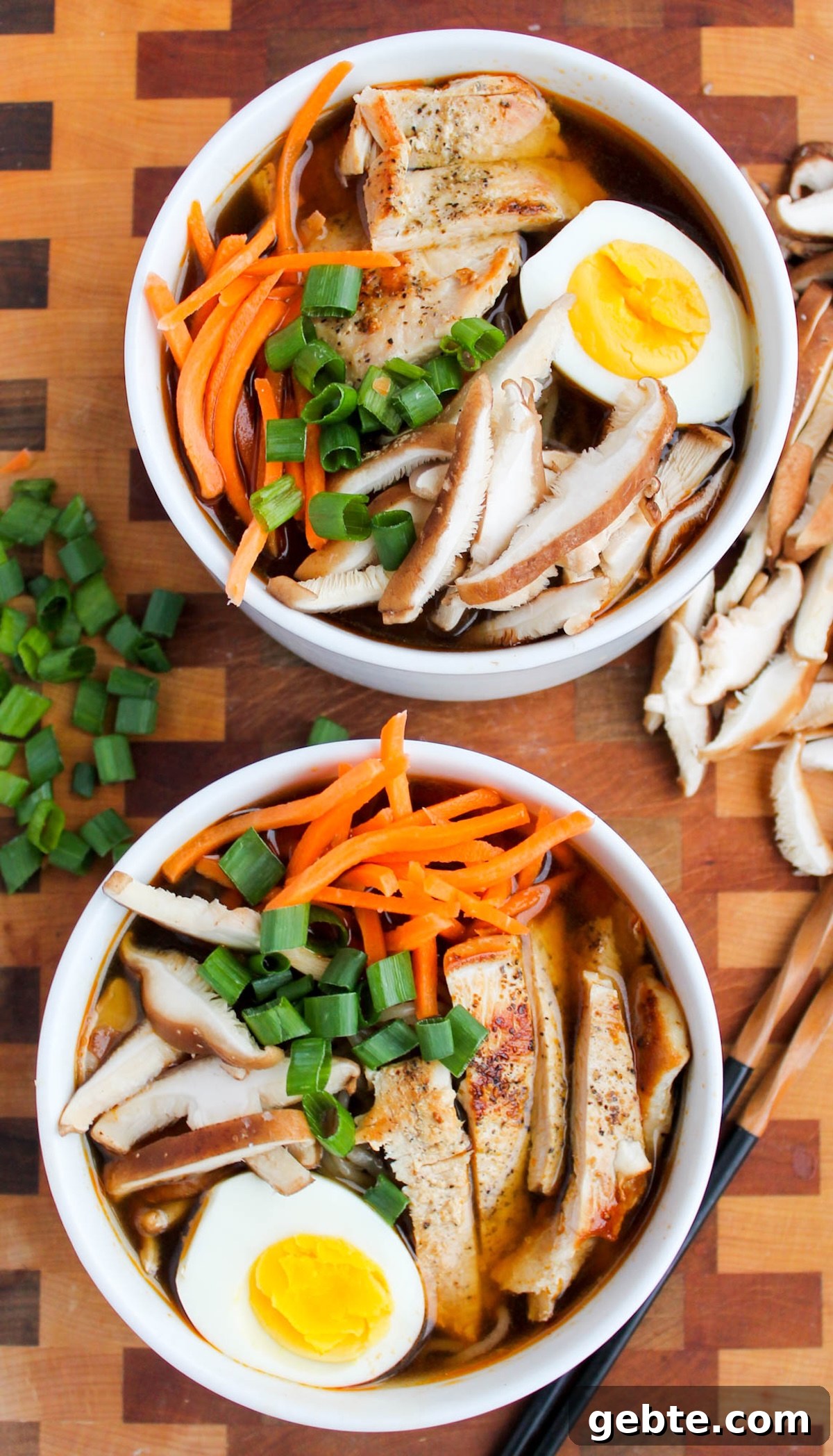 Two white soup bowls each filled with a homemade ramen that's topped with a half hard-boiled eggs, shredded carrots, slide chicken breast, and sliced mushrooms.