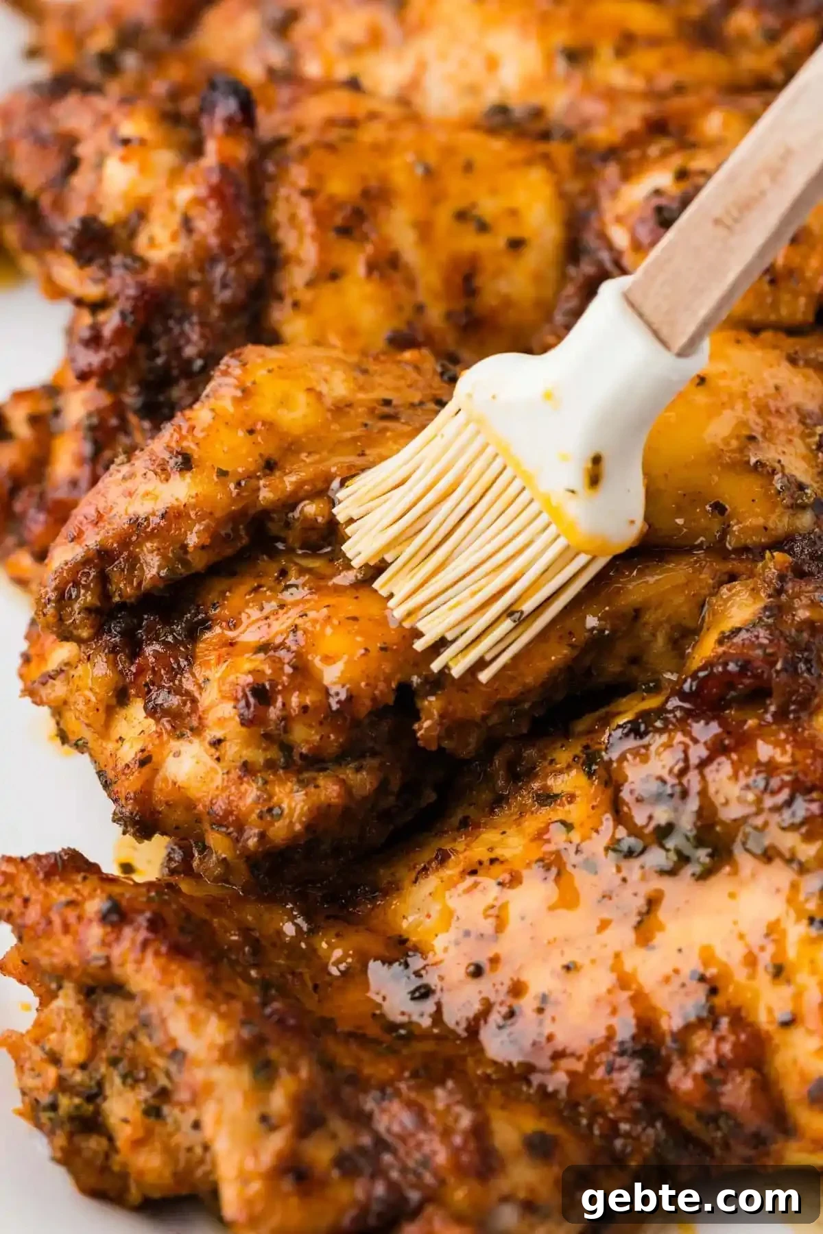 A chicken thigh coated in a reddish yellow marinade is being brushed by a silicone pastry brush. Several other chicken thighs are out of focus in the background.