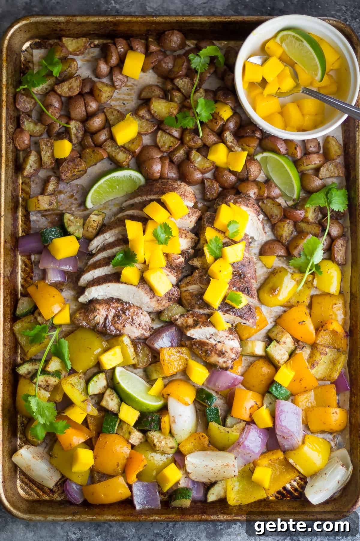 A sheet pan is filled with sliced chicken breast that's coated in a dark marinade, chopped yellow peppers, red onion, and potatoes. A small bowl of diced mango and a lime wedge sits on the side.