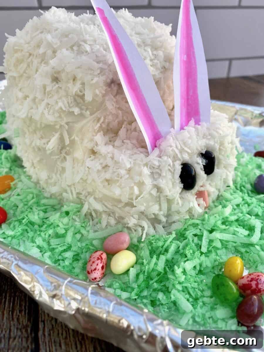 Bunny cake from the side with large paper ears standing up straight and jelly beans and tinted coconut on the side.