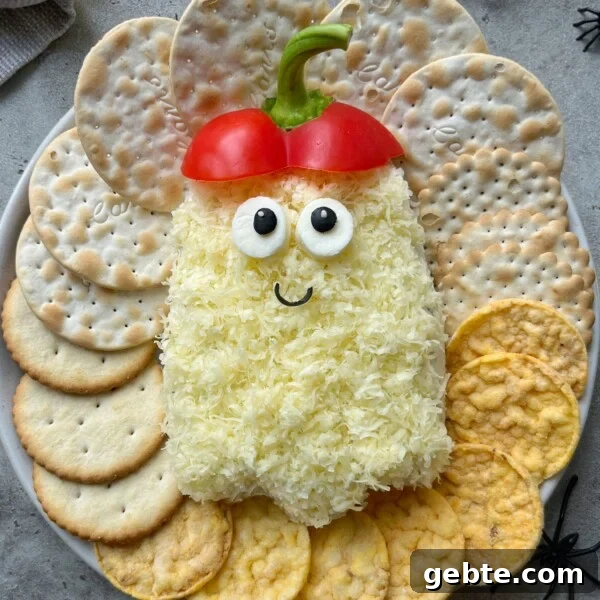 A Halloween-themed cheese ball shaped like a ghost, with shredded cheddar cheese, olive eyes, red bell pepper hat, and a smiling face. Surrounding it are assorted crackers.