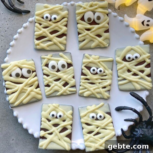 A plate of rectangular chocolate pieces decorated as mummies with white chocolate bandages and candy eyes. Plastic spider and ghost decorations surround the plate.
