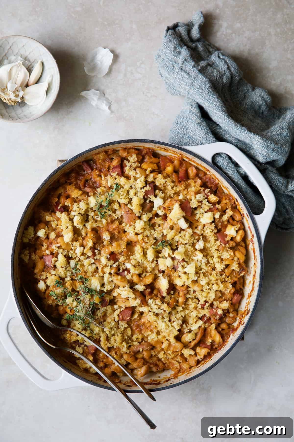 Photo of Easy Weeknight Chicken and Sausage Cassoulet with serving utensils with blue napkin and garlic cloves.