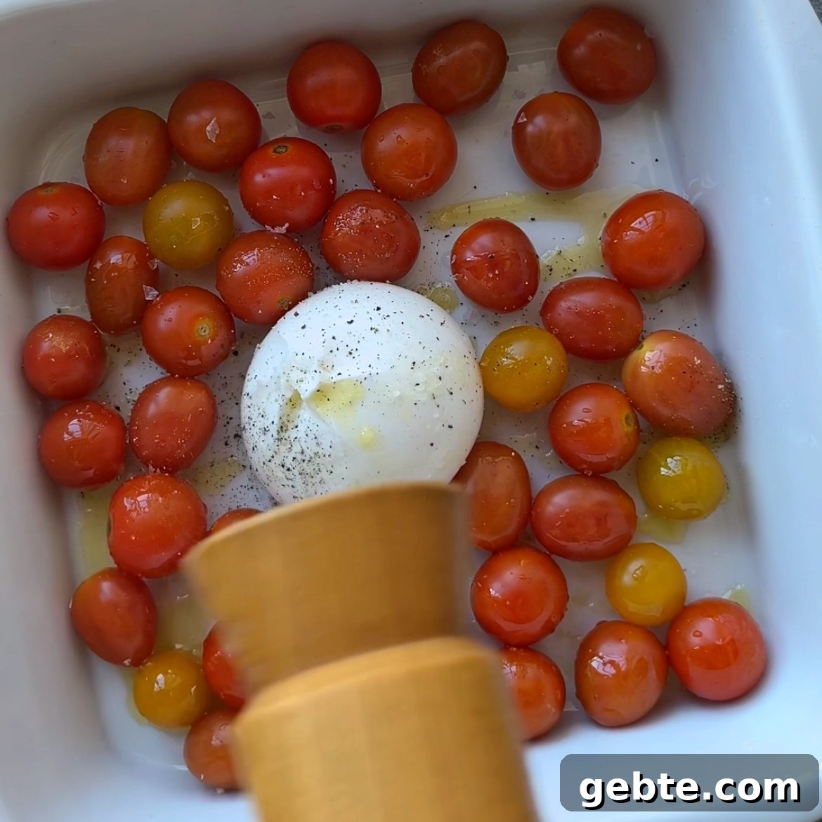 Creamy Burrata Pasta with Roasted Cherry Tomatoes 3 A white baking dish with cherry tomatoes and a ball of burrata cheese is being seasoned with freshly ground pepper.