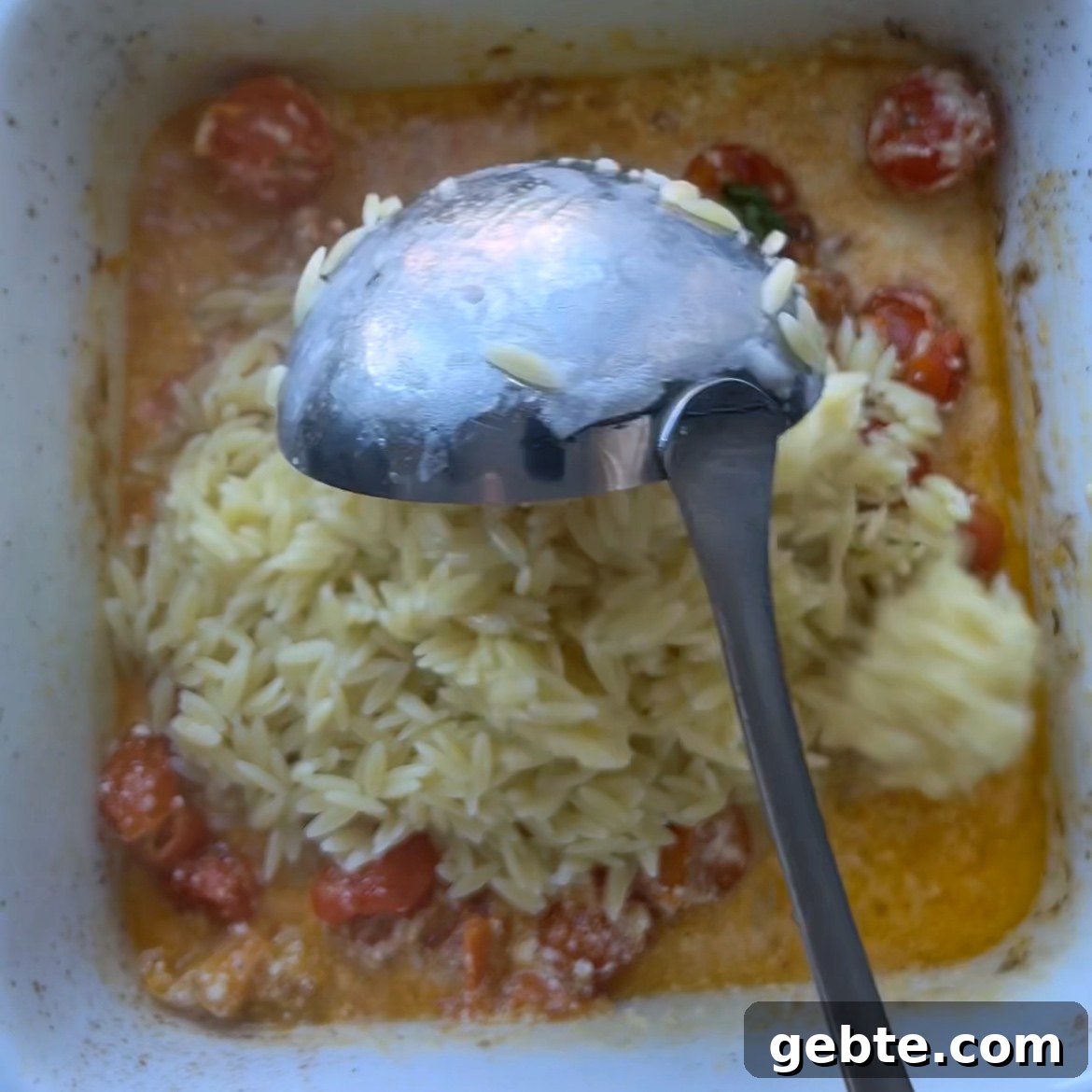 Creamy Burrata Pasta with Roasted Cherry Tomatoes 5 A metal spoon stirs pasta, cherry tomatoes, creamy sauce, and rich burrata cheese in a square white baking dish.