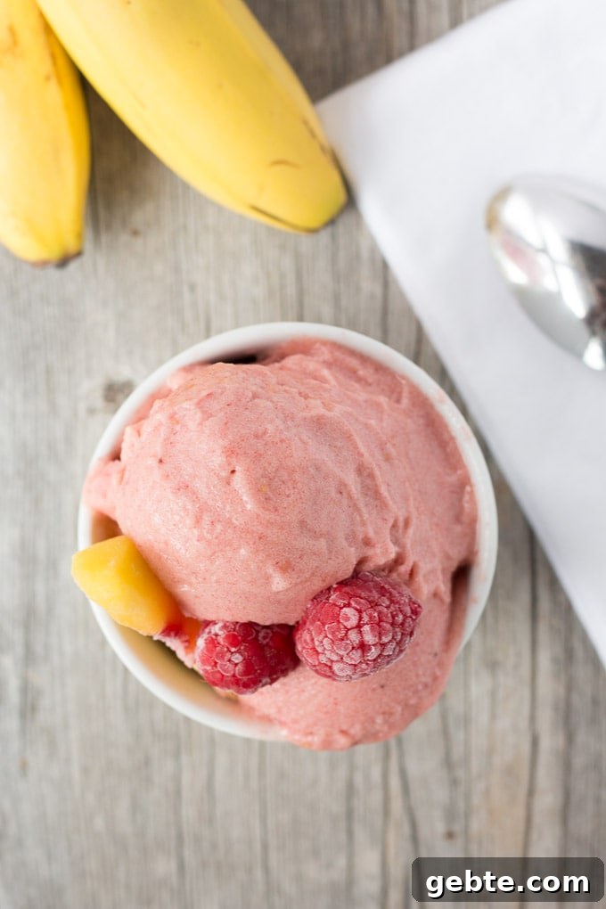 Top down shot of a pink nice cream with frozen raspberries and a peach slice all in a small white bowl on top of a gray background.