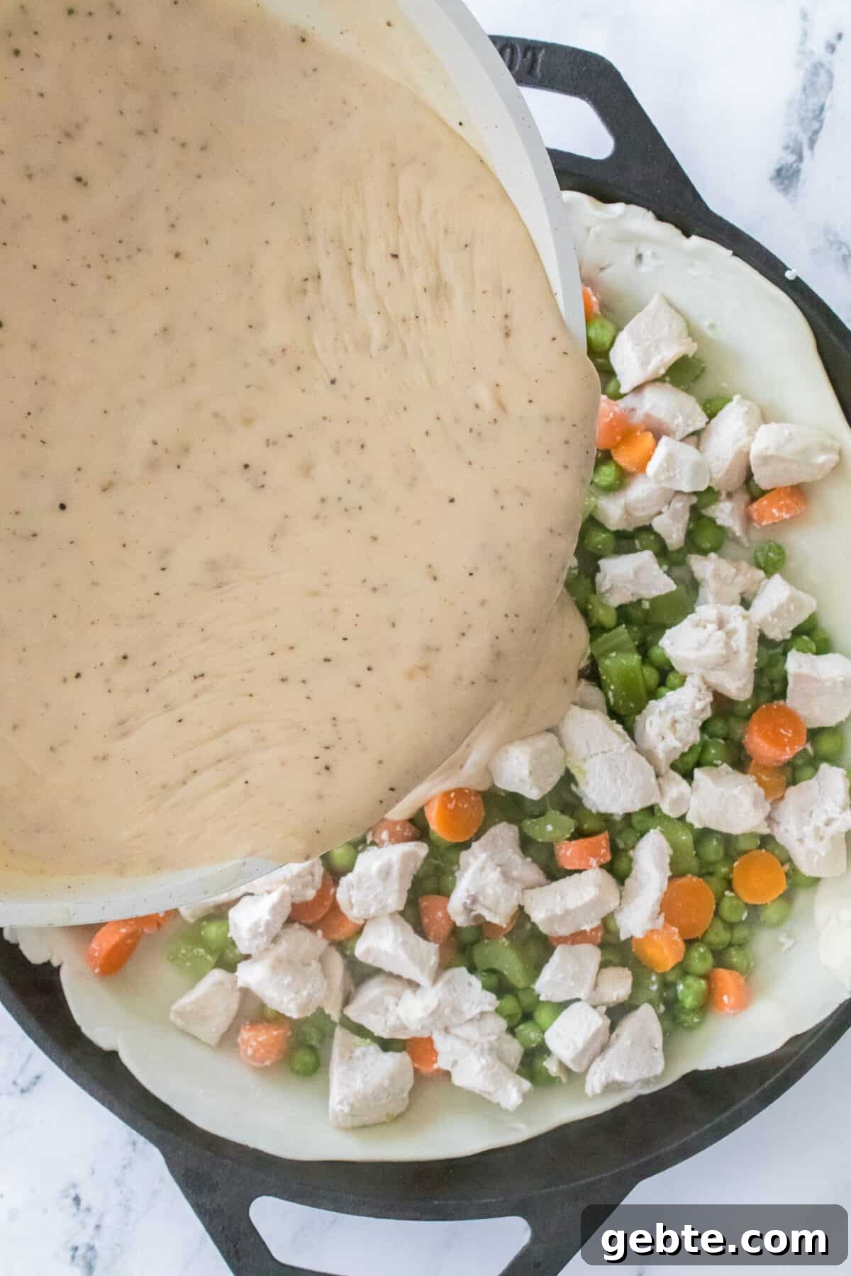 overhead shot of creamy roux being poured over cast iron skillet with pie crust, chicken, peas, celery and carrots