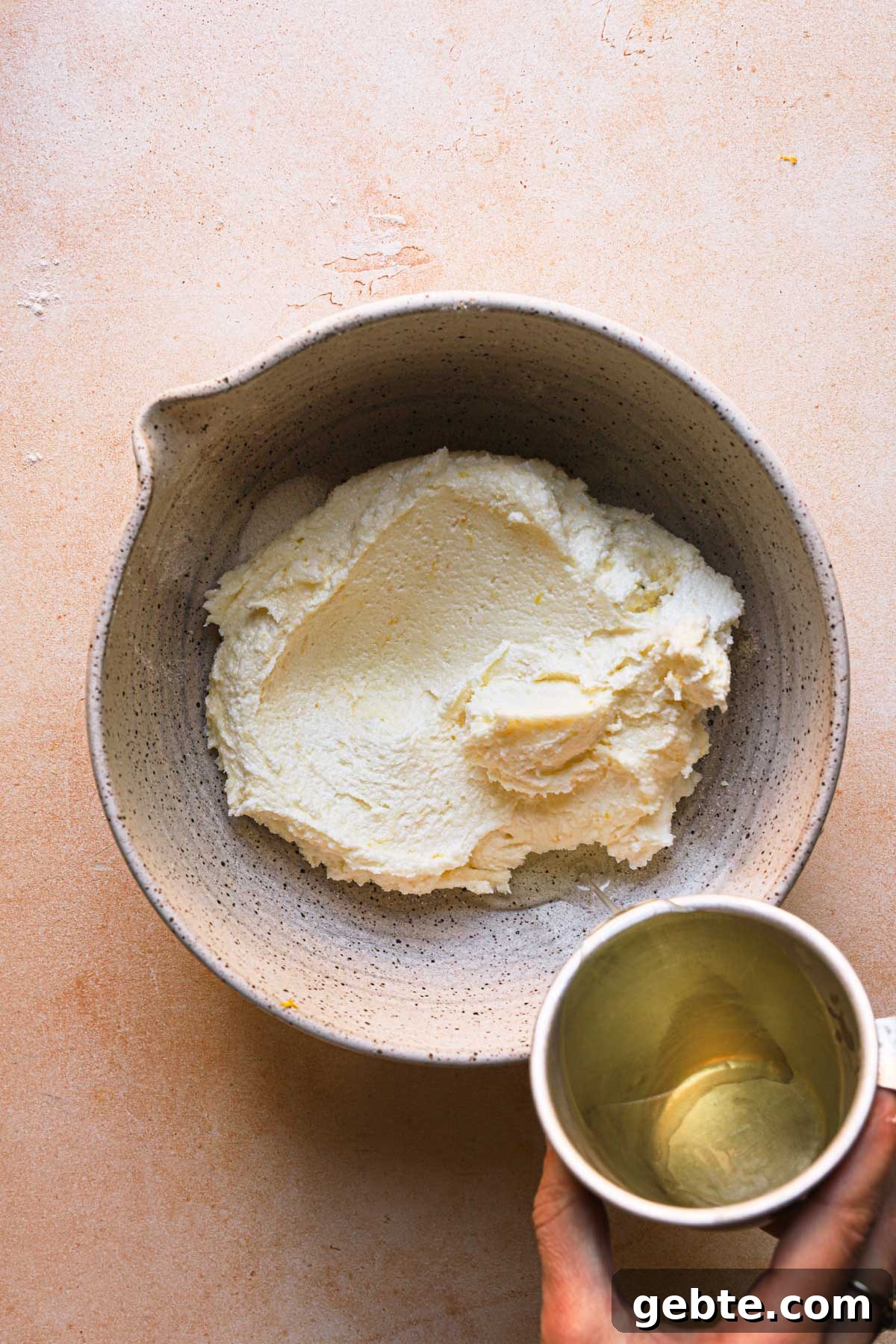 A hand pouring olive oil into a bowl with creamed sugar and butter.