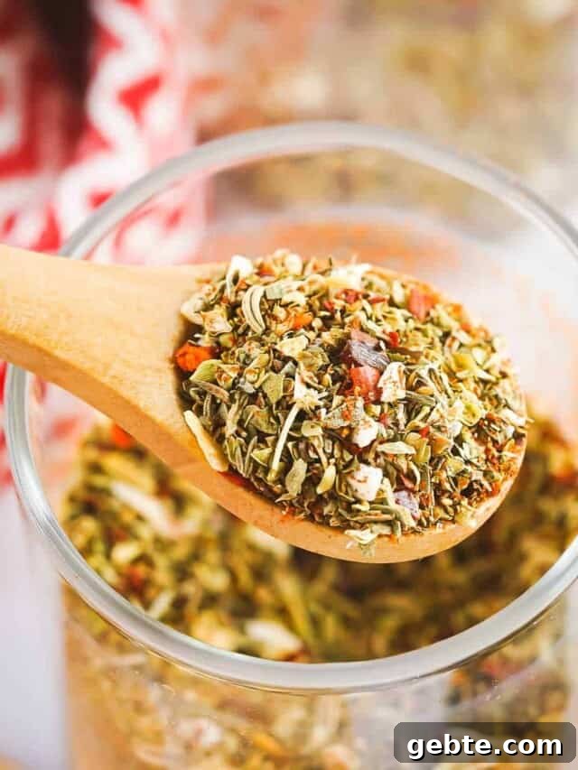 Close up shot of a wooden spoon holding a scoop of pizza spices over glass jar full of them with a red cloth in the background.