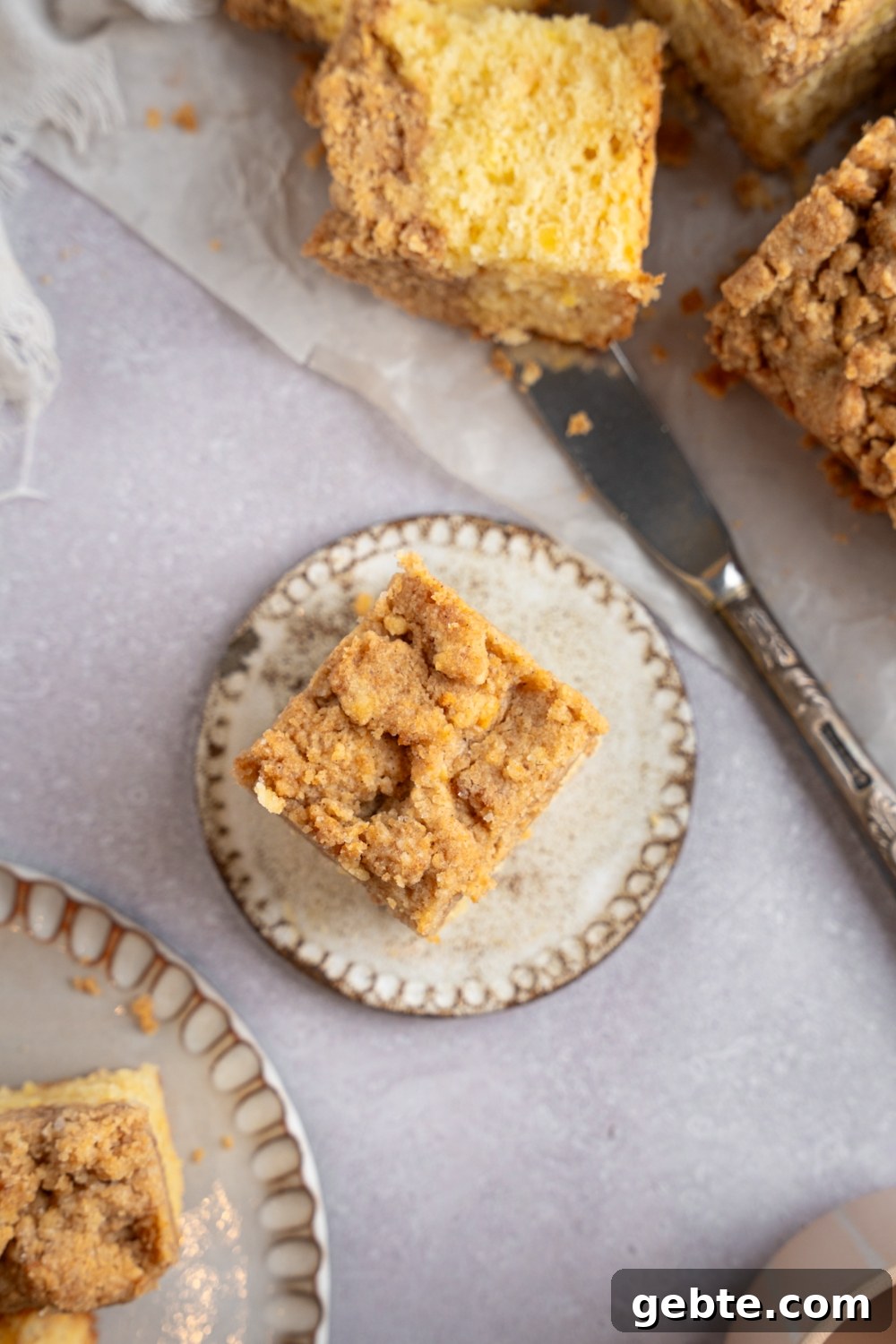 A square slice of Bisquick coffee cake on a ceramic plate. The plate is on a white counter with additional slices of cake on a sheet of parchment paper. There is a knife next to the coffee cake.