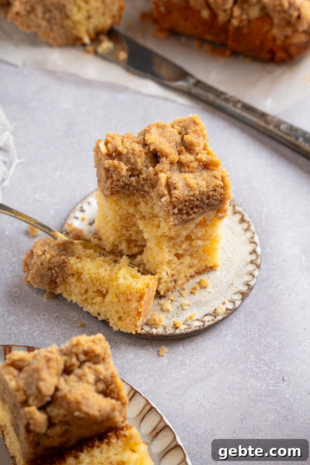 A square slice of Bisquick coffee cake on a ceramic plate with a fork slicing a bite. The plate is on a white counter with an additional plate of coffee cake.