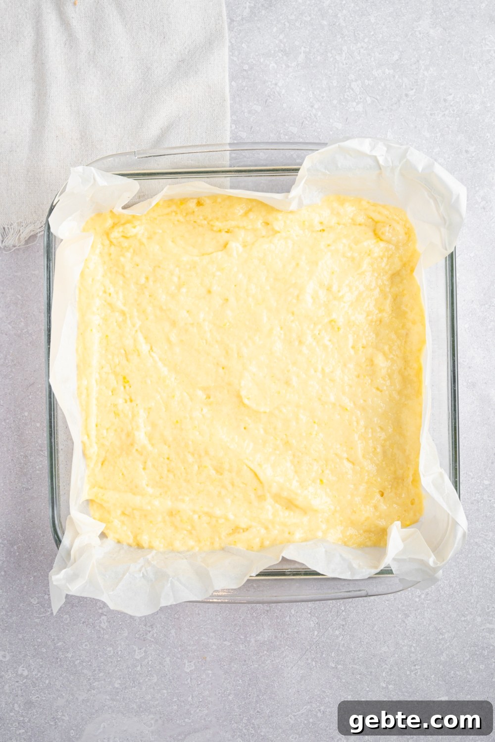 A square glass baking dish lined with parchment paper and filled with Bisquick dough. The dish is on a white counter with a white cloth napkin.